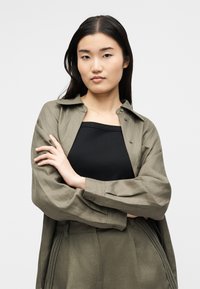 Γυναίκα με μακριά μαύρα μαλλιά φοράει πουκάμισο και παντελόνι ελιάς πάνω από μαύρο τοπ, με σταυρωμένα χέρια, κοιτάζει μπροστά με ουδέτερη έκφραση.