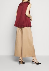 Top senza maniche in satins color bordeaux con schiena a occhiello, abbinato a pantaloni beige a gamba larga e scarpe nere con tacco alto. Tessuti lisci e vestibilità comoda.
