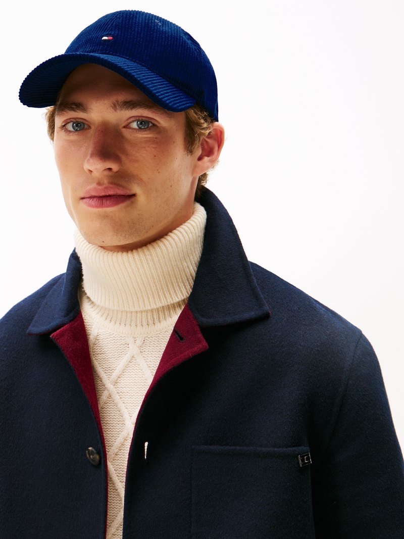 Cappotto di lana navy con dettagli bordeaux, indossato sopra un dolcevita di maglia color crema. Cappello in corduroy blu con logo. Strati caldi e dall'aspetto strutturato evidenti.
