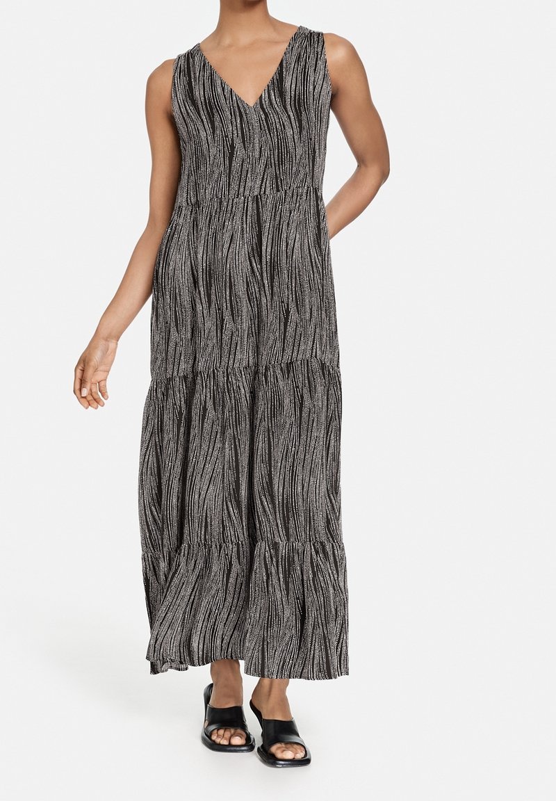 Robe longue sans manches à motif noir et blanc avec col en V portée avec des sandales noires à bout ouvert, une main du mannequin posée sur la hanche.