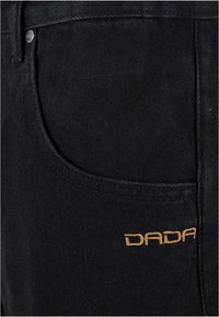 Melni denima džinsi ar mazliet teksturētu virsmu, ar sānu kabatu un oranžu izšuvumu ar vārdu "DADA" priekšā.