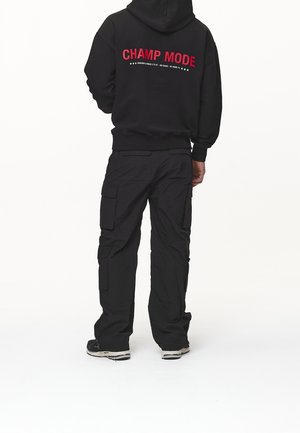 Mann trägt schwarzen Kapuzenpullover mit rotem "CHAMP MODE" Schriftzug auf dem Rücken und schwarze Cargohosen, steht vor weißem Hintergrund.