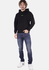 Felpa nera con cappuccio e tasca frontale, con logo verde. Abbinata a jeans blu e sneakers bianche con dettagli neri.