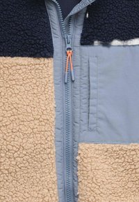 Giacca in pile con pannelli navy, crema e azzurro chiaro; presenta una cerniera centrale con dettagli arancioni e una tasca laterale.