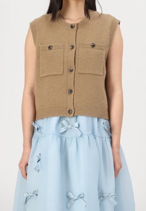 Ärmellose braune Strickweste mit Knöpfen und zwei Fronttaschen, getragen über einem hellblauen Rock, der mit kleinen Stoffschleifen verziert ist.