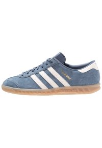 Blue suede sneaker with three white stripes, gold branding, and light brown rubber sole. Low-top design and rounded toe shape.