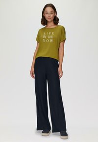 Olijfgroen t-shirt met witte tekst "LEEF IN HET MOMENT", gecombineerd met donkere wijde broek en zwarte sneakers met dikke zolen.