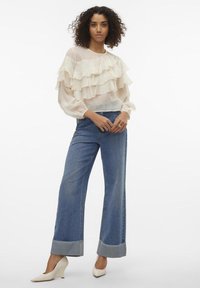Blouse blanche à volants superposés associée à un jean en denim bleu à jambes larges avec des poignets repliés. Le mannequin porte des chaussures pointues de couleur crème.