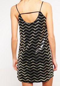 Kobieta w czarnej i srebrnej cekinowej mini sukience w wzór chevron bez rękawów, z cienkimi ramiączkami, widoczna z tyłu.