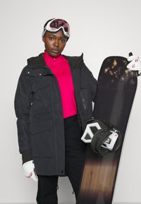 Black snowboard jacket with multiple pockets, pink zip-up layer underneath, black pants, white gloves, and a snowboard with a dark, gradient design.