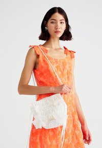 Bolsa branca texturizada com detalhes florais, alça em corrente e laços. Modelo vestindo um vestido laranja vivo com um padrão floral.