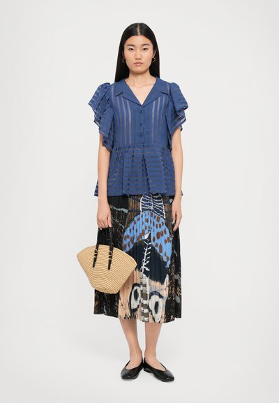 Woman wearing blue striped short-sleeve blouse, abstract patterned midi skirt, black flats, and holding beige woven handbag.
