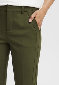 Olivroheline kohandatud pükste mudel küljetaskutega, siledast tekstuurist ja kitsast lõikega. Omab sirgeid jalgu koos peente õmblustega.