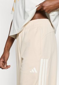 Bézs sportos melegítőnadrág rugalmas derékpánttal, oldal zsebbel és fehér Adidas logóval, három csíkkal díszítve.