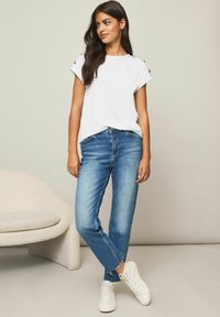 Hvid kortærmet top med knapdetaljer på skuldrene, parret med lyseblå denimjeans med afslappet pasform og frynsede kanter. Hvide sneakers.