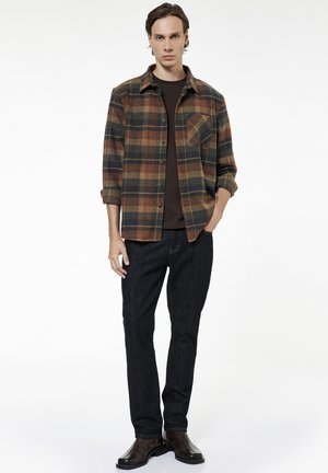 Uomo con camicia a quadri marrone, verde e arancione sopra una maglietta nera, jeans scuri e stivali di pelle marrone, in piedi davanti a uno sfondo bianco.