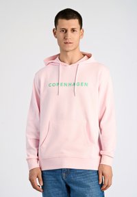 Ljusrosa hoodie med känguruficka, med grön text "KÖPENHAMN". Tillverkad av mjukt tyg, den har en huva med dragsko och ribbade ärmslut.