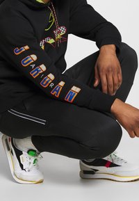 Sudadera negra con un gráfico multicolor en el frente y "JORDAN" impreso en naranja y azul en la manga. Pantalones negros con rayas blancas.