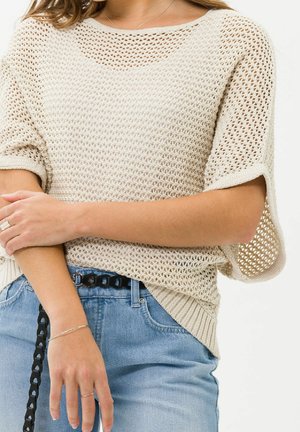 Kobieta w beżowym, luźnym, ażurowym swetrze z krótkim rękawem, zestawionym z jasnoniebieskimi dżinsami i czarnym plecionym paskiem.