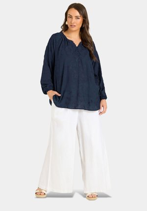 Donna che indossa una blusa larga blu scuro a maniche lunghe con motivi floreali e pantaloni bianchi ampi, in piedi con una mano in tasca.