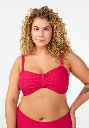 Kučeravá žena v červenom bikini vrchu s nastaviteľnými ramienkami, stojaca s jednou rukou na bokoch proti jednoliatemu bielemu pozadiu.