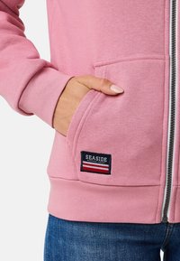 Rosa Zip-Hoodie mit weicher Textur, Kängurutasche und einem dunkelblauen "SEASIDE" Logo-Patch mit roten und weißen Streifen.