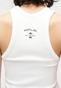 Vue arrière d'une personne portant un débardeur blanc côtelé avec l'inscription "REPLAY NOT ORDINARY" en noir sur le haut du dos.