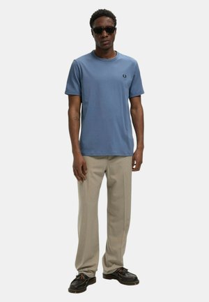 Uomo con occhiali da sole, t-shirt blu a maniche corte, pantaloni beige e scarpe nere, in piedi su uno sfondo bianco uniforme.