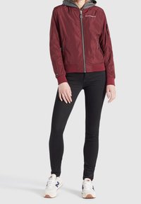 Burgundská bomber bunda se šedou kapucí, vybavená předním zipem, žebrovanými manžetami a kapsou na levém rukávu. Černé úzké džíny a bílé tenisky.