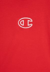 Sarkans kokvilnas T-kreklīgs ar mazu baltu "C" logo izšūtu kreisajā krūtī. Gluda tekstūra ar īsām piedurknēm.