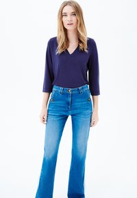 Meripaita tummansinisellä V-päällä ja kolmella neljänneksellä hihoilla, yhdistetty korkeavyötäröisiin, leveälahkeisiin sinisiin farkkuihin, joissa on kultaisia yksityiskohtia taskuissa. Tasainen kankaan rakenne.