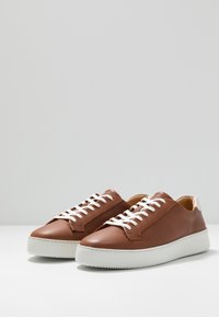 Bruna lädersneakers med vita snören, rund tå och texturerad vit gummisula. Minimalistisk design med en slät yta och diskret sömnad.
