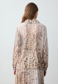 Woman with long brown hair wearing a beige and brown snake print long-sleeve shirt and matching pleated skirt, facing away against a plain background.