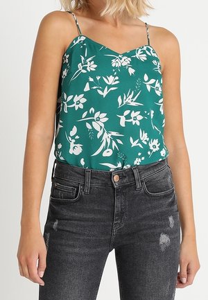 Vrouw draagt een groene mouwloze top met bloemenpatroon, gecombineerd met hooggesloten, vervaagde zwarte jeans met lichte slijtage.
