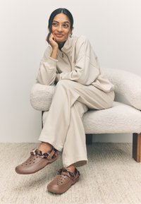 Beige zip-up sweatshirt and matching wide-leg pants, paired with brown clogs featuring a fur trim and perforated design, seated on a textured chair.