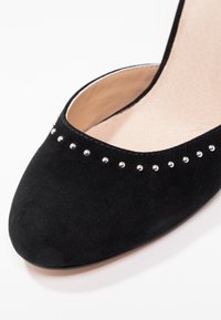 Svart mocka högklackad sko med rund tå, prydd med silvriga nitar längs kanten och en strukturerad innerfoder.