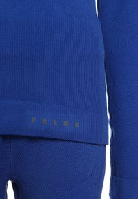 Blaues Strickkleidungsstück mit gerippter Textur, das das Logo "FALKE" in Gold aufweist. Das Design umfasst einen eng anliegenden, schichtweise aufgebauten Stil für Wärme.
