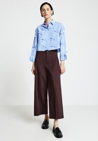 Donna che indossa una blusa corta azzurro chiaro con piccoli fiocchi e pantaloni marroni a gamba larga e a vita alta, in piedi davanti a uno sfondo bianco.