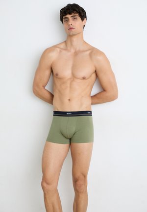Grüne Baumwoll-Boxershorts mit einem schwarzen Bund, auf dem "BOSS" in Weiß steht. Sie haben eine enge Passform und eine glatte Textur. Das Modell steht.