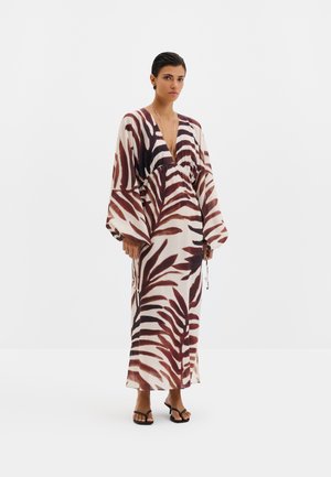 Femme debout dans une longue robe ample aux manches larges, présentant un motif abstrait de feuilles marron et blanc, portant des talons noirs ouverts à l'avant.