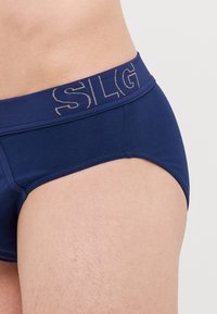 Navyblaues Herrenbikini mit einem weichen elastischen Bund, der goldene Schriftzüge trägt. Hergestellt aus einem glatten Material mit einem schlanken, enganliegenden Design.