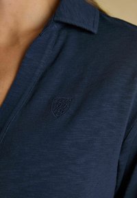 Lähedalne pilt inimesest, kes kannab tume sinist struktureeritud särki, millel on rinnal väike kirjatud kilbi logo.