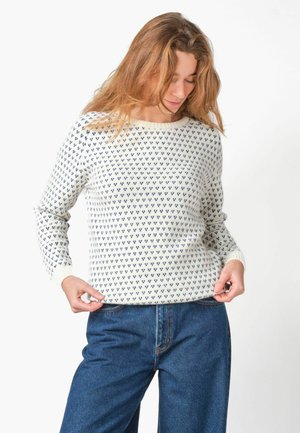 Vrouw met lang haar die omlaag kijkt, gekleed in een witte trui met kleine blauwe patronen en een blauwe spijkerbroek, en de zoom van de trui met beide handen vasthoudt.