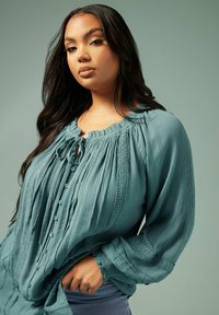 Teal, plisserad blus med rund halsringning, justerbart band och delikata spetsdetaljer. Knappdetaljer och långa ärmar med rynkade manschetter.