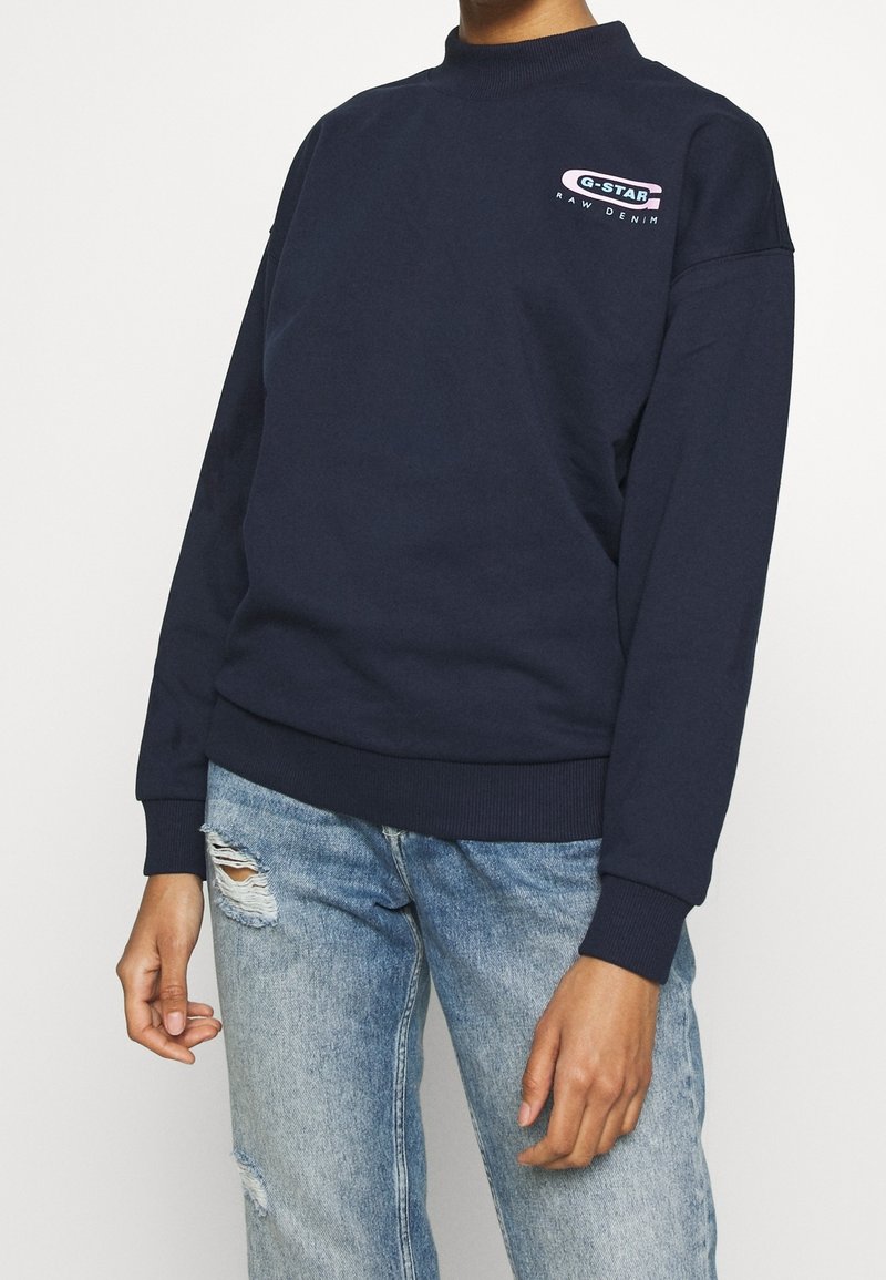 Mörkblå sweatshirt med hög hals, ribbade manschetter och fåll. Har en vit och rosa logotyp på vänster bröst. Bärs tillsammans med slitna ljusa jeans.
