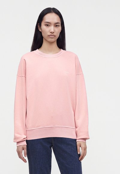 Jeune femme aux longs cheveux foncés portant un sweat-shirt rose clair et un jean bleu foncé, debout devant un fond clair uni.