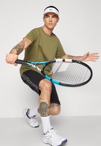 Homem atlético com uma t-shirt verde-azeitona e calções pretos, segurando uma raquete de ténis com um design azul e preto. Ténis brancos e viseira.