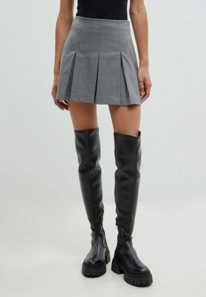 Woman wearing a gray pleated mini skirt and black over-the-knee chunky platform boots, standing against a plain background.