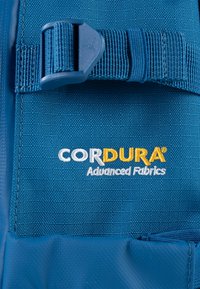 Modra Cordura tkanina z teksturirano površino; vsebuje rumeni in beli logotip "CORDURA® Advanced Fabrics" ter podrobnost s plastično sponko.
