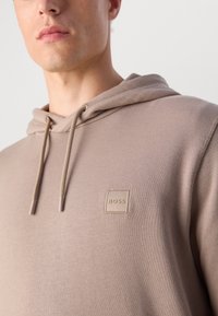 Man in een beige hoodie met een trekkoord en een klein vierkant BOSS-logo op de borst, uitgesneden onder de neus en boven het midden van de torso.